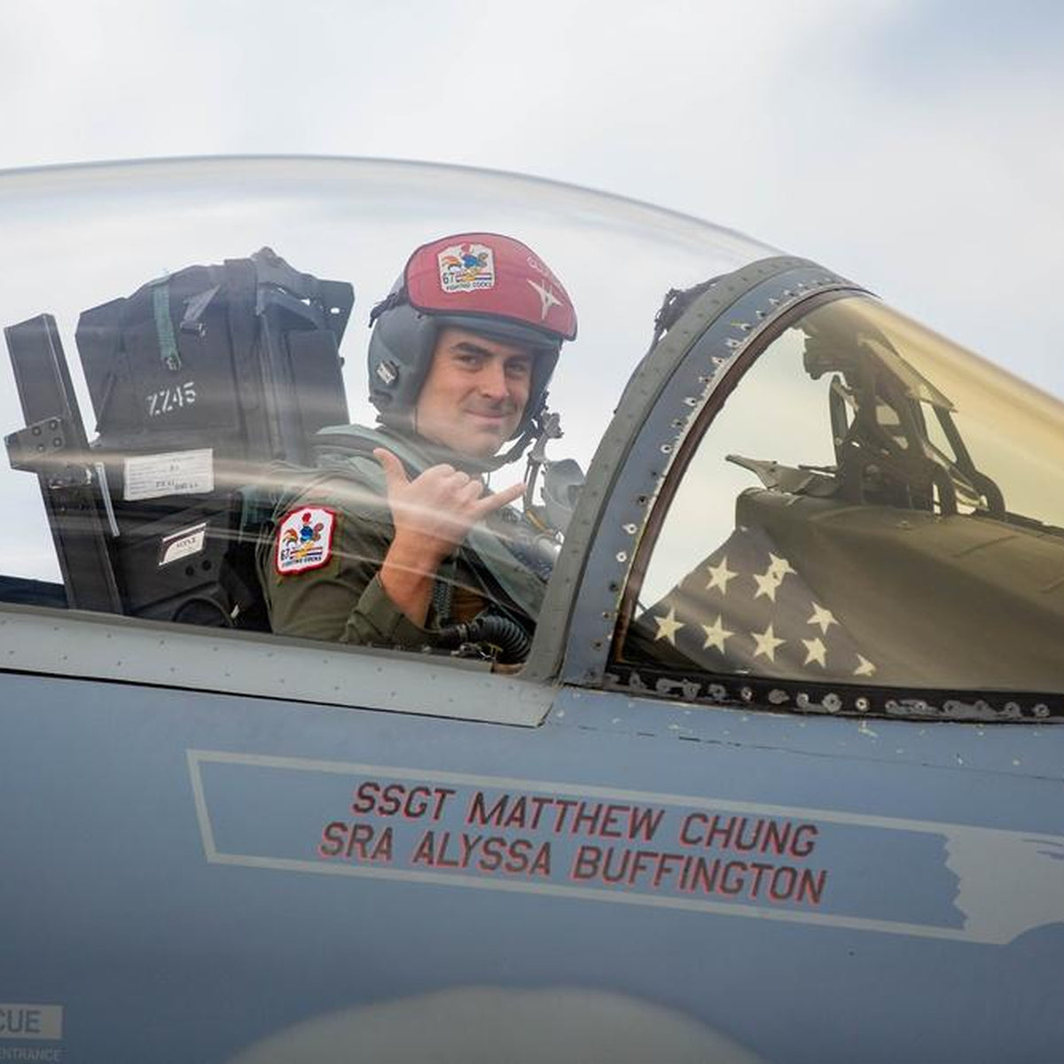 A pilot wearing Fighting Cocks patches makes a shaka gesture in an F-15 cockpit beside a folded American flag. Crew names “SSGT MATTHEW CHUNG” and “SRA ALYSSA BUFFINGTON” appear below.