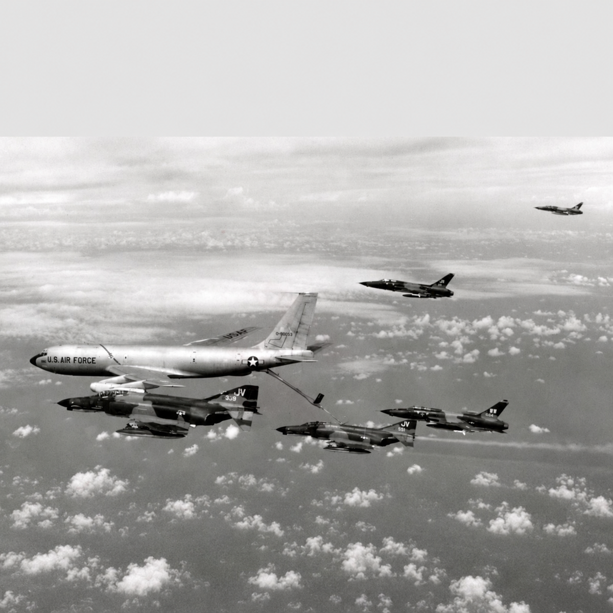 Black-and-white photograph of a KC-135 Stratotanker refueling an F-4 Phantom II above the clouds, with other F-4s flying nearby.