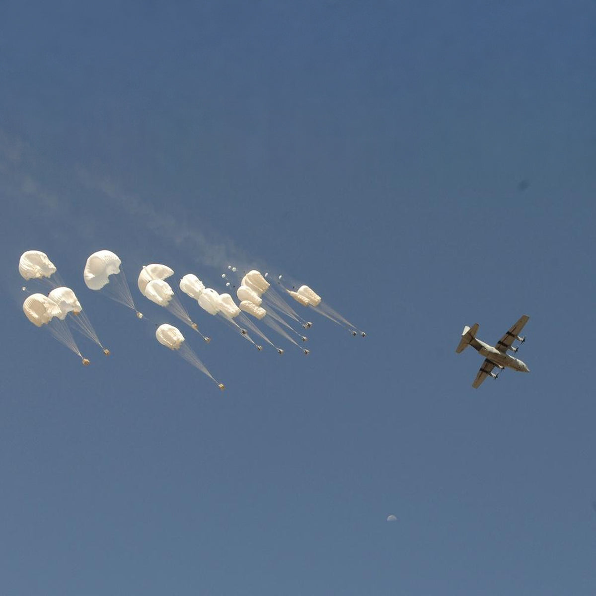 C-130 Hercules flies past supply bundles descending beneath white parachutes, with the moon visible below.