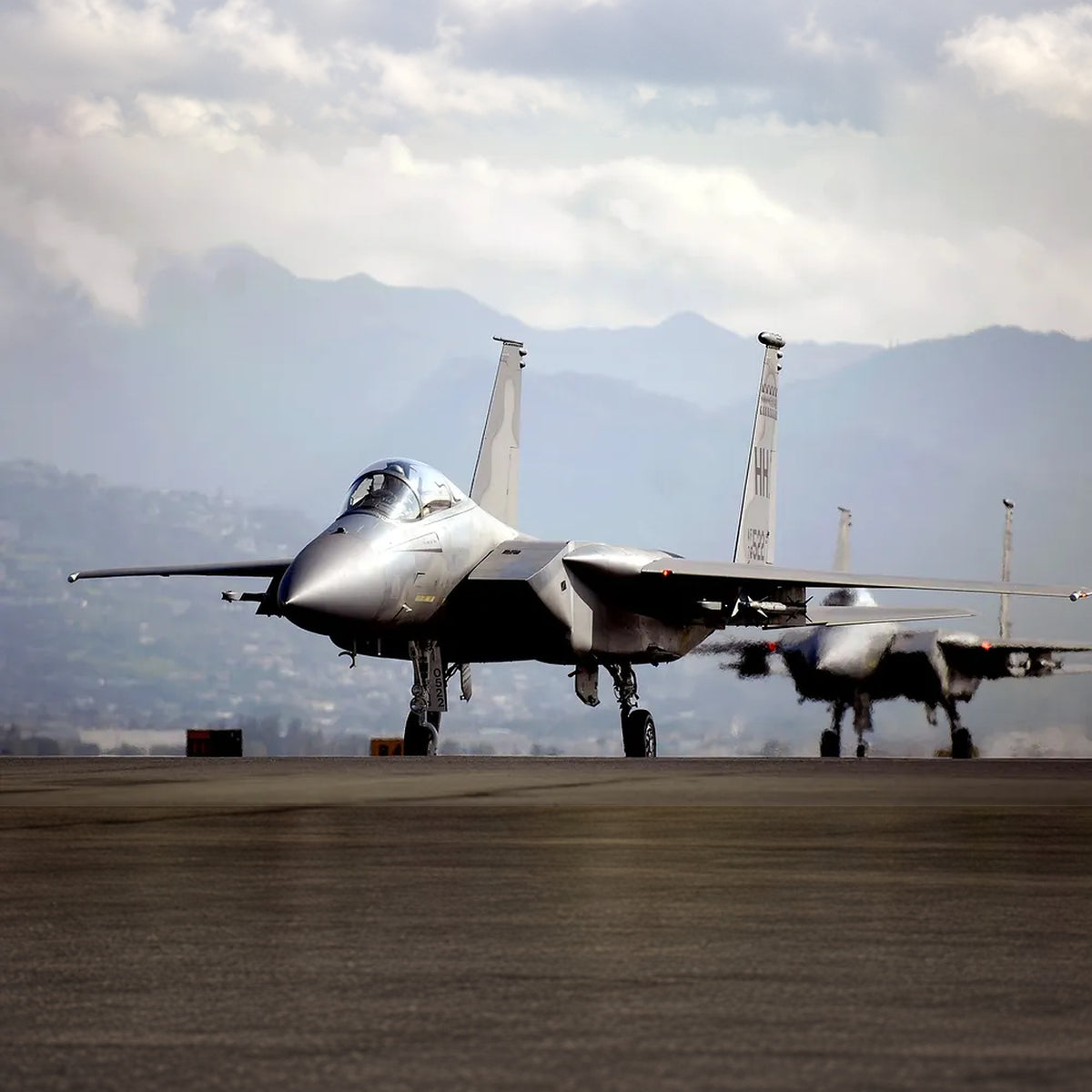 Two F-15A Eagles on an airfield, viewed from the front with landing gear down, twin tail fins, and mountains beneath a cloudy sky.