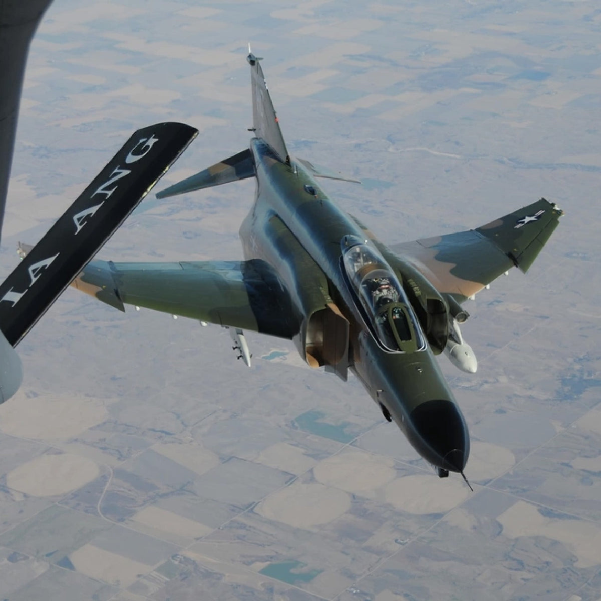 Camouflaged F-4 Phantom II approaching an aerial refueling boom above patchwork farmland, with its two-seat cockpit and swept wings visible.