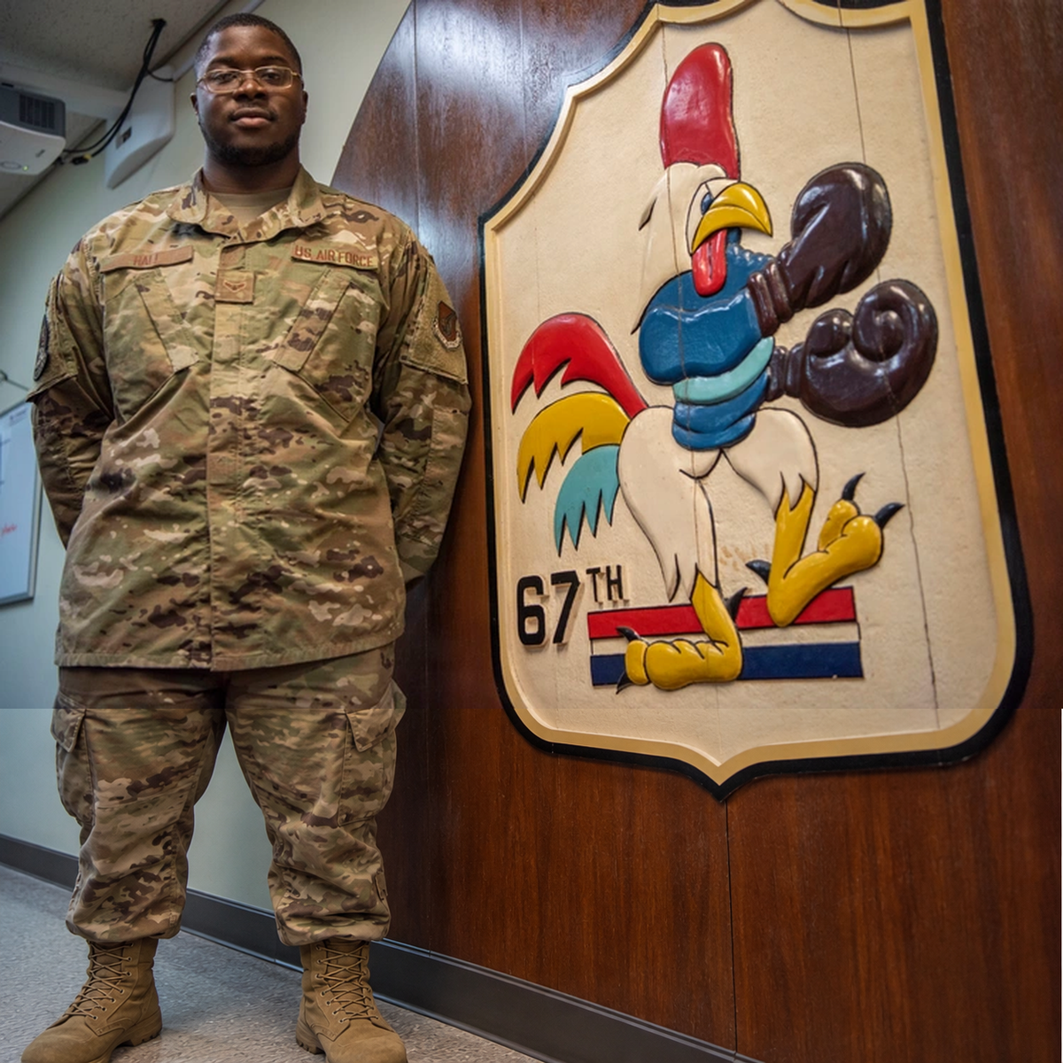 Airman in camouflage beside a large “67TH” insignia depicting a boxing rooster.