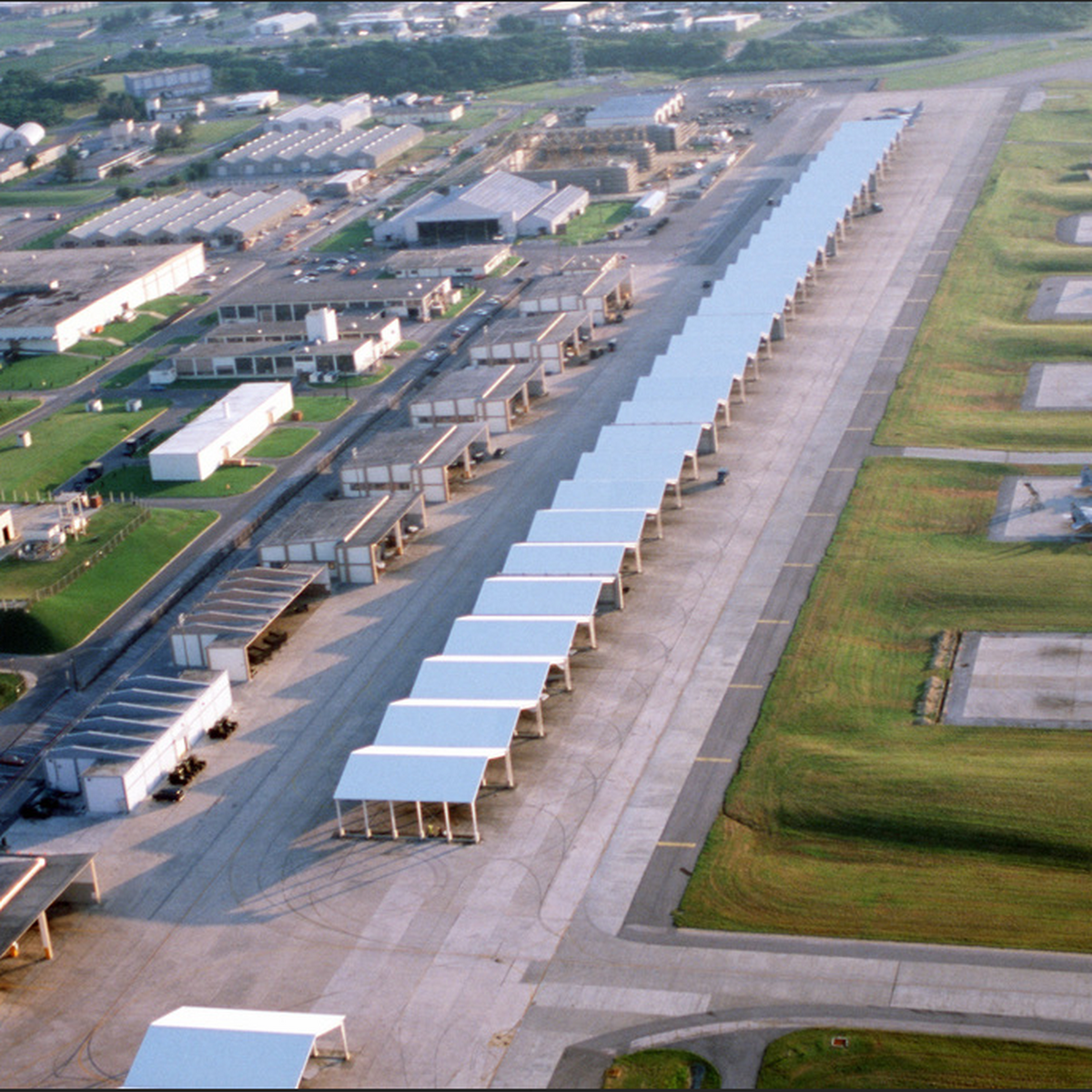 Aerial view of Kadena Air Base in Okinawa, Japan, showing long rows of covered aircraft parking shelters beside taxiways, grassy areas, and base buildings.