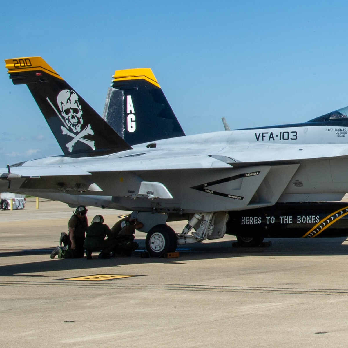 White skull and crossbones on a VFA-103 Super Hornet’s black-and-yellow tail, with sailors beneath the aircraft and “HERE’S TO THE BONES” lettering.